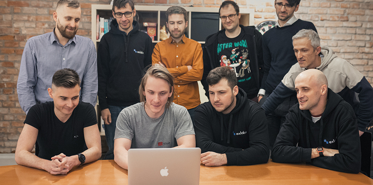 Group of nine men gathered around a laptop on a wooden table in a casual office setting with a brick wall background.