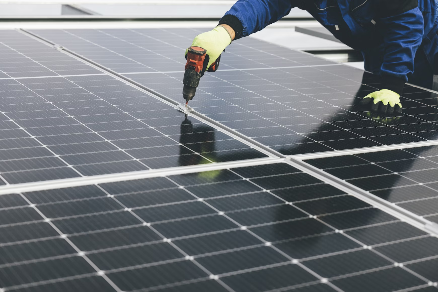 Worker in gloves installing solar panels using a power drill on a rooftop.