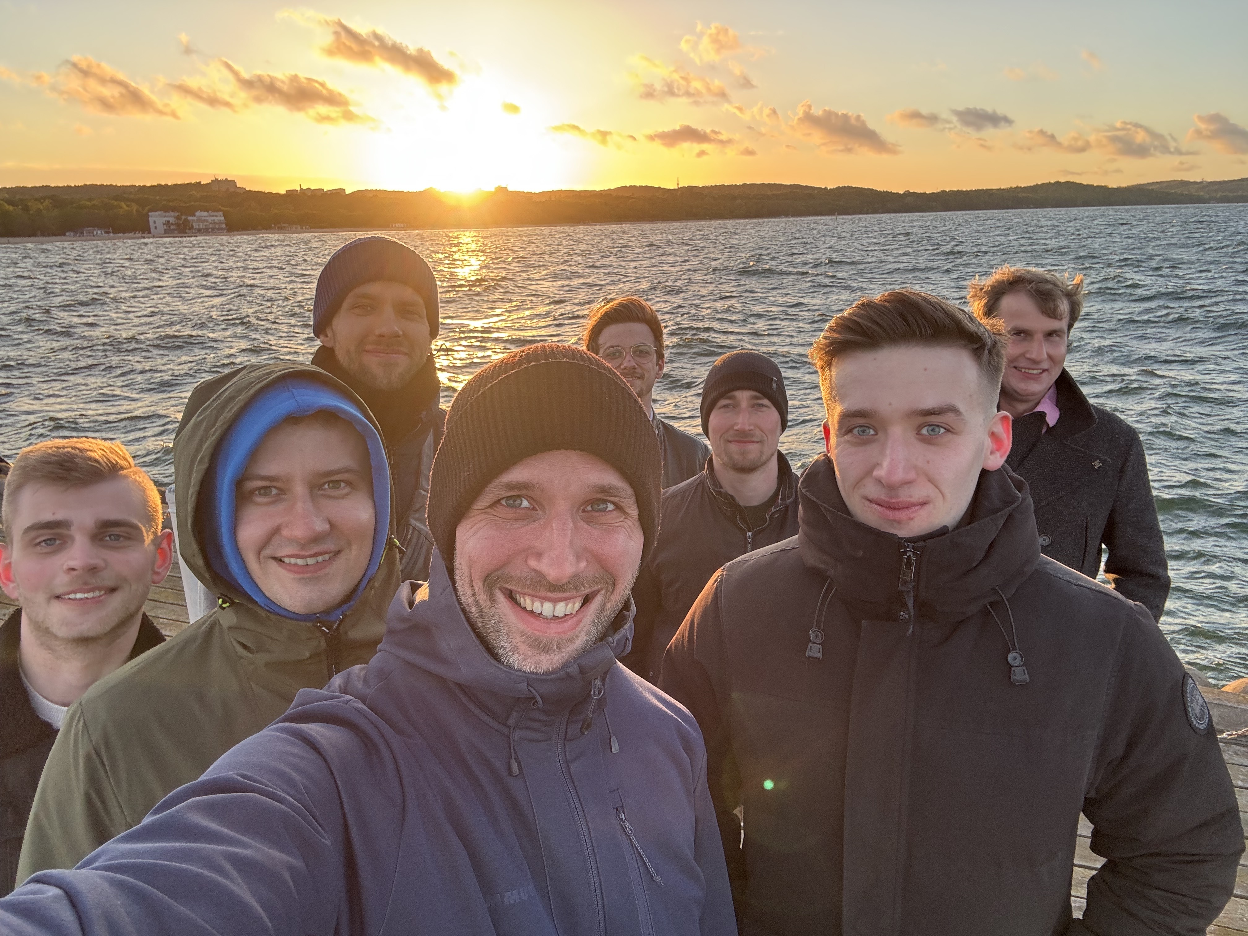 Group of eight men in jackets and hats smiling at the camera on a pier with a sunset over a lake in the background.