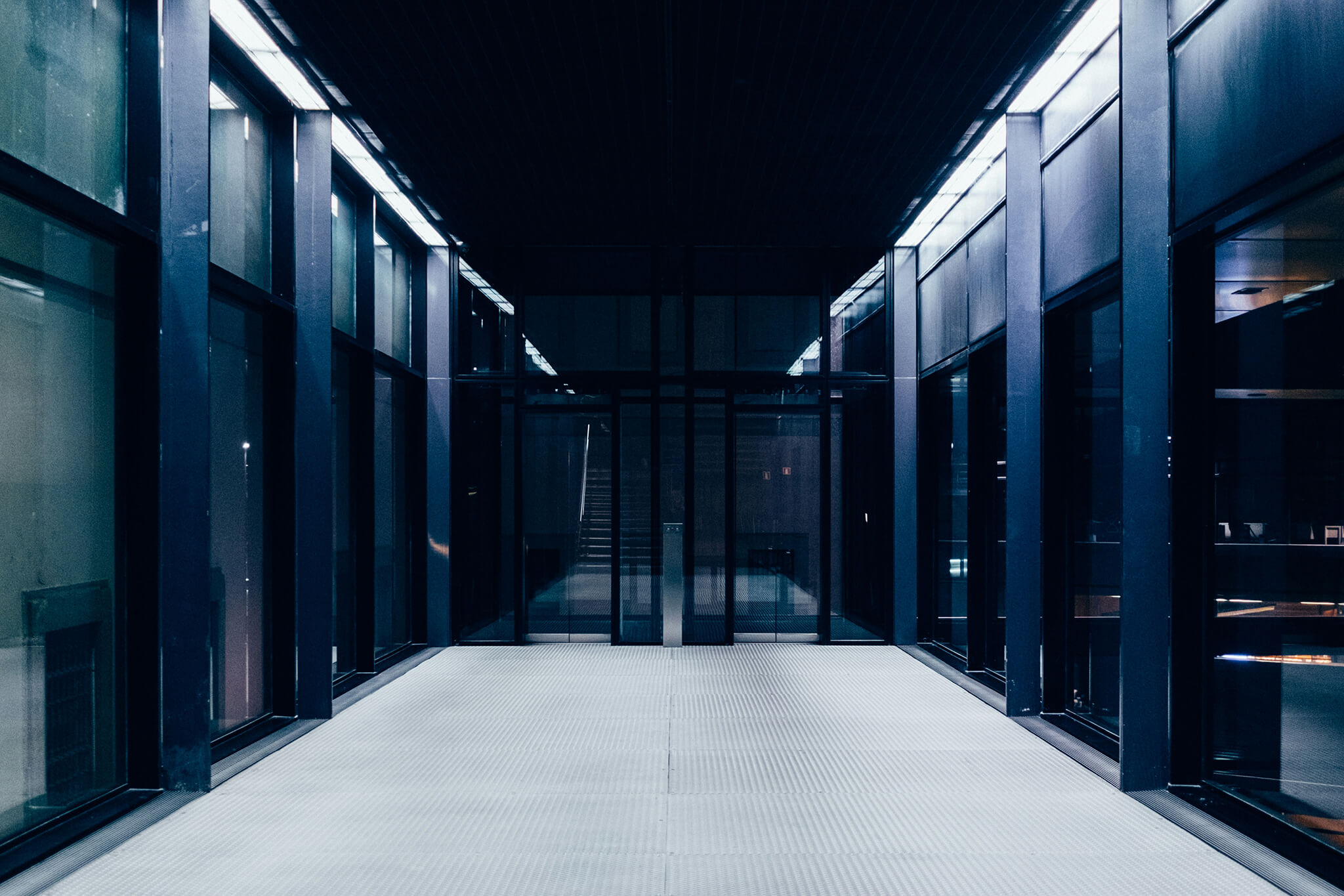 Empty corridor with glass walls and a metal grated floor leading to dark glass doors, illuminated by overhead lights.